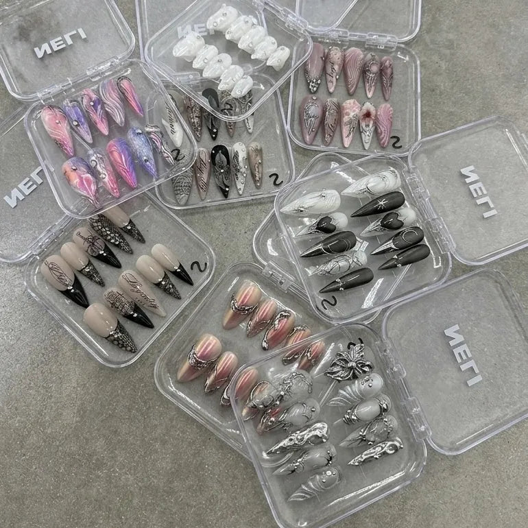 Sets de press-on nails NELI dans leurs boîtes transparentes sur un fond gris.