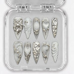 Ongles press-on NELI argentés et blancs avec ornements en relief, présentés dans une boîte transparente.