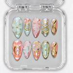 Ongles press-on NELI corail et nacrés avec fleurs en relief et ornements dorés, dans une boîte transparente.